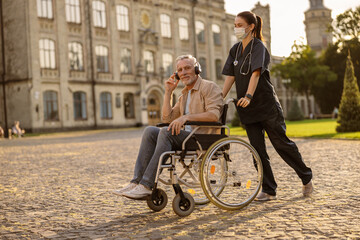 Fototapeta na wymiar Young female care taker in protective mask spending time with senior man in wheelchair wearing headphones and listening to music outdoors in the park near clinic