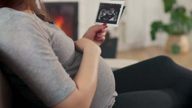 Beautiful pregnant woman with brown hair stitting in front of cosy fireplace stroking baby belly look at ultrasound pictures shot in 4k