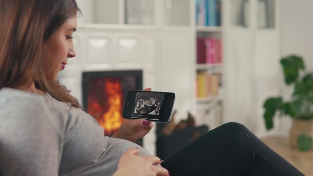 Beautiful pregnant woman with brown hair stitting in front of cosy fireplace stroking baby belly look at ultrasound pictures on smartphone shot in 4k