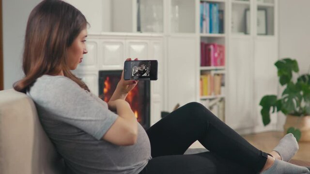 Beautiful pregnant woman with brown hair stitting in front of cosy fireplace stroking baby belly look at ultrasound pictures on smartphone shot in 4k