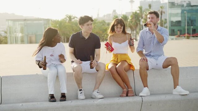 Four multiracial people best friends listening to music and drinking beer on embankment at sunset