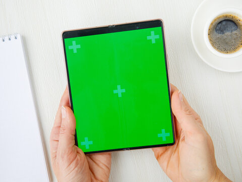 woman's hand holding new folding Smartphone Samsung Galaxy Z Fold 2 with green screen, open notebook, cup of coffee lying on white table. Flat lay top view, mock up. October4,2021 Novosibirsk,Russia