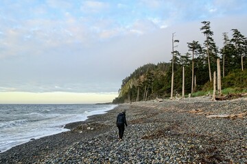 A woman walking along agate beach with the waves crashing to shore and the sun setting, with tow hill in the background, on a beautiful evening on Agate Beach, Haida Gwaii, British Columbia, Canada