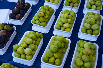 Fresh green figs neatly arranged in white trays on a blue tablecloth at an outdoor market. The figs are plump, ripe, and full of natural sweetness, with some darker figs visible in the background. 