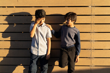Caring for sad friend. Two young boys standing resolving conflict. Giving help concept. Kid offering support. Golden hour scene of upset child with hand on face. Hand on shoulder.