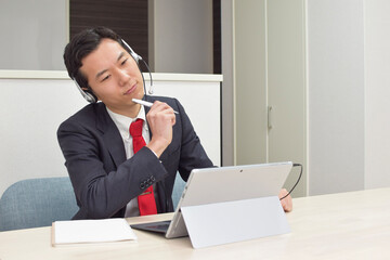 A young Asian man wearing a headset thinks about how to deal with his customer during telework at home