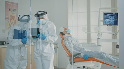 Stomatologists consulting digital tablet for patient oral care in orthodontic cabinet. Caucasian medical team of dentists using technology and modern equipment while wearing ppe suits
