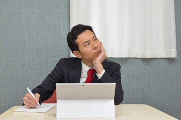 A young Asian man wearing business suit thinks about new idea and writes some memos on his notebook during telework at home