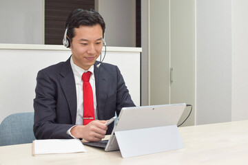 A young Asian man wearing a headset points a pen to his pc screen during telework