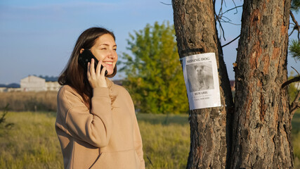 Smiling woman in hoodie dials number from missing dog poster hanging on tree trunk to inform owners...
