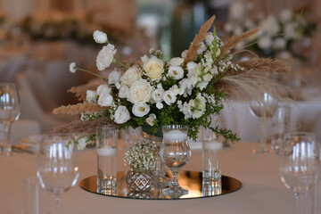 White tablecloths with clear vases and white flowers and fern arrangements. Golden colored plates, peavh napkins, table numbers and mirror centerpieces.