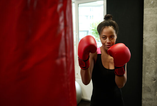 Beautiful African fit woman with perfect physique posing at camera wearing red boxing gloves, hitting punching bag in gym. Female boxer training hard during martial combat art