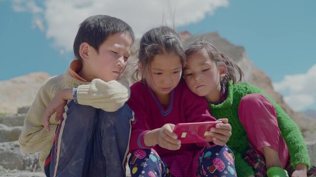 Three little adorable Asian village kids sitting outdoors and engrossed in watching a movie or video using mobile phone sitting in a mountainous region during daytime. concept of distant connectivity
