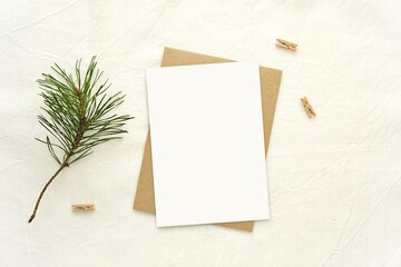 Christmas greeting card mockup, blank vertical A6 card, brown envelope, pine branch and wooden pins, linen table cloth  background, minimal aesthetic style.