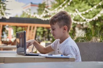 little schoolboy sits outdoors, types on keyboard of laptop. smiling caucasian boy sitting at table at veranda of cafe, studying with computer, doing homework. distant learning, online education