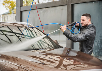 Guy washing dirt from windshield of his auto, using powerful stream of water. Rinsing machine by...