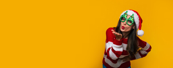 Banner wide photo of Crazy and excited beautiful modern woman in funny Christmas glasses and Santa hat is having fun while posing against a yellow wall