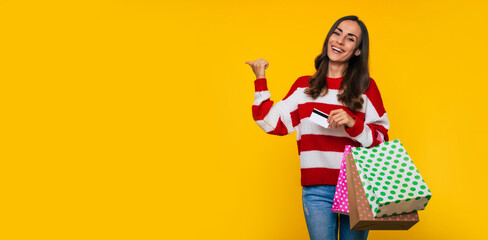 Banner photo of beautiful young happy and excited woman in a striped sweater with many colorful shopping bags and credit card in hands isolated on yellow background