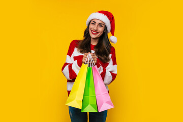 Happy stylish beautiful woman in Christmas hat with colorful shopping bags isolated on yellow background
