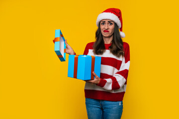 Young disappointed cute girl in Christmas hat is unpacking gift box on yellow background with...