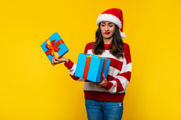 Young disappointed cute girl in Christmas hat is unpacking gift box on yellow background with...