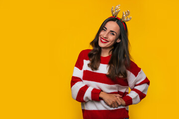 Beautiful funny young woman in a striped winter sweater and deer hat on the head is posing isolated on yellow background in studio