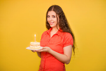 Portrait of a girl in a red shirt with a piece of cake and candle on a white plate in her hands isolated on a yellow background.