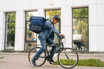 Side view of handsome young delivery man in protective helmet starting bike ride in city street on background of office building. Courier male with thermo backpack delivery food to client