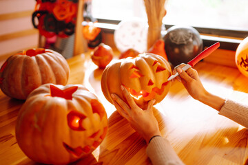 Close up of woman hands with knife carving pumpkin. On the table lies a orange pumpkin with a painted horrible face. Halloween, decoration and holidays concept.