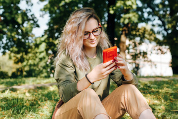 Young hipster woman in glasses using mobile phone while sitting on green grass outdoors. Smiling teenage girl chatting, sending messages, surfing internet or social media on smartphone in nature