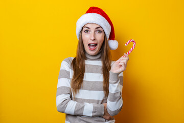 Fototapeta na wymiar Surprised young woman in Santa Claus hat with Christmas candy on a yellow background