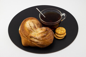 Still life with yeast sweet heart shaped bun with a cup of black tea and crispy crackers on a black tray and white background. Russian tea drinking concept. Top view