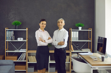 Serious young woman and elderly man in white shirts standing arms folded in modern office. Portrait of two confident people, business professionals, company owners and co founders with arms crossed - obrazy, fototapety, plakaty