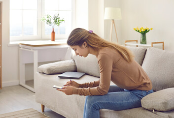 Woman using modern mobile phone at home. Serious young girl sitting on sofa with laptop computer,...