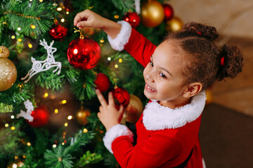 A portrait of black girl in a red dress decorates a Christmas tree with a glass red ball at home.