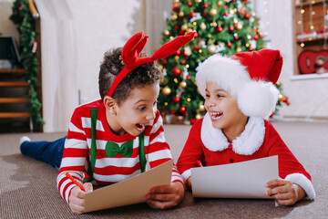 A black boy and a girl lie on the floor near a decorated Christmas tree and read letters for Santa