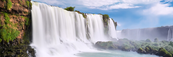 Naklejka premium Iguazu waterfalls in Argentina, view from Devil's Mouth. Panoramic view of many majestic powerful water cascades with mist. Panoramic image with blue sky and clouds.