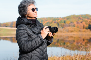 Elderly woman photographer standing against of autumn forest and lake in nature. Side view of...