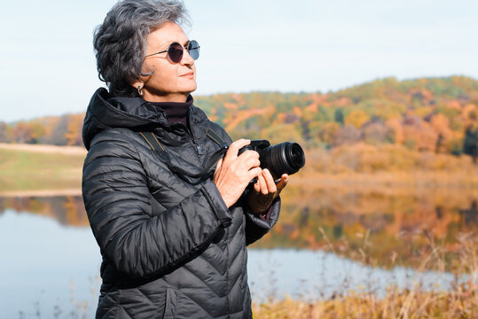 Elderly woman photographer standing against of autumn forest and lake in nature. Side view of senior woman with glasses using digital camera for landscape photo outdoors. Seniors and Technology