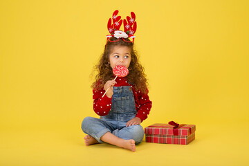 Little girl for the Christmas holidays with a gift and a lollipop on a yellow background to copy  space
