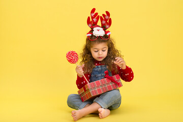 Little girl for the Christmas holidays with a gift and a lollipop on a yellow background to copy  space