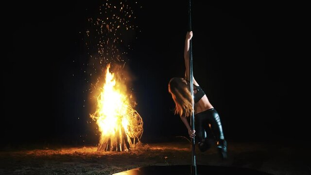 pole dance. witch halloween. fit female gymnast, in black leather outfit and high heels, performs acrobatic exercises on metal rotating pole, at night, by light of large fire in background.
