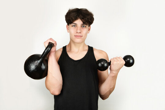The teenager guy on a light background holds a heavy kettlebell and a dumbbell, a guy of strong physique and with a beautiful body, he is engaged with kettlebells and dumbbells for health