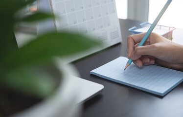 Close-up of a business woman hand writing notes on paper notepad, making agenda on personal organizer with desktop calendar and laptop computer on office table at workplace