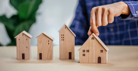 Businessman choosing mini wood house model from model and row of coin money on wood table, selective focus, Planning to buy property. Choose what's the best.