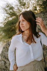 portrait of a young woman with dark straight hair in a white shirt against the background of nature. summer, sunset sun, authentic smiling brenette