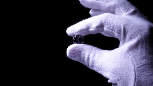 Goldsmith hands in white gloves inspect the quality of luxury brilliant diamond. Selective focus, macro. Precious stone shine in the light with star rays. Round diamond for Engagement or wedding ring