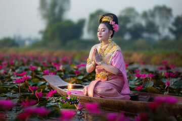Beautiful Thai woman in Thai traditional costume on the wooden boat and holding floating basket (Krathong in Thai language) for floating in the red lotus lake in Loy Krathong Festival of Thailand.