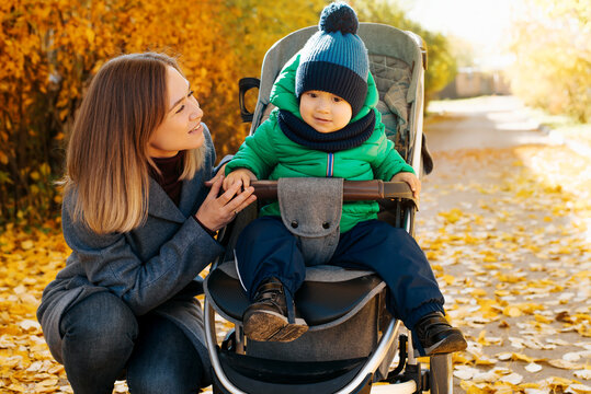 Family portrait mom and little son in baby carriage in autumn outdoors. Smiling young mother looking at child on walk in nature. Motherhood, parenting concept