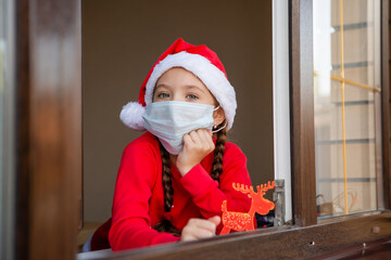 Fototapeta na wymiar New Year and Christmas are quarantined. Stay at home. A girl in a medical mask looks out of window. winter concept. space for text. High quality photo